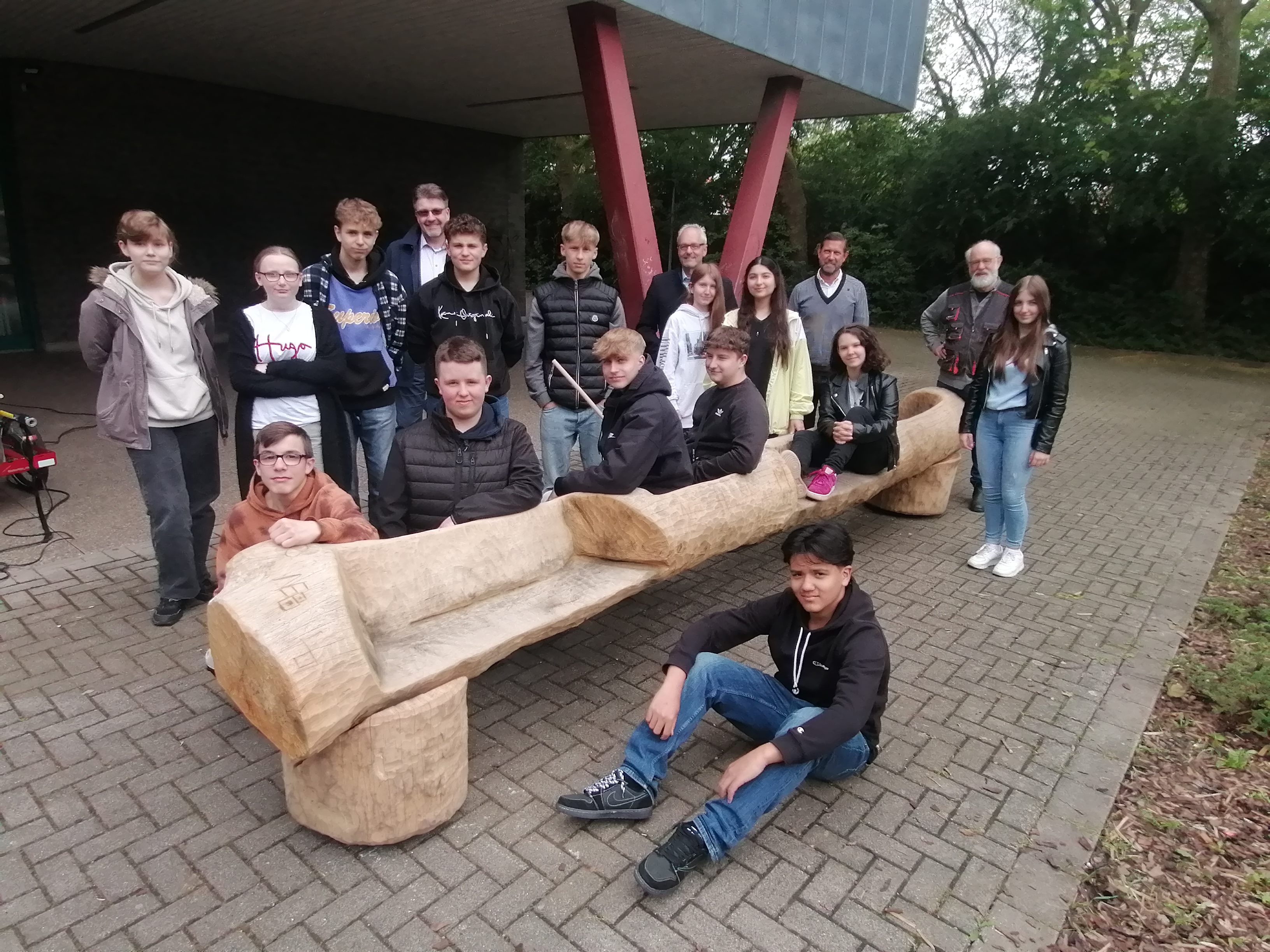 Schülerinnen und Schüler der AG Holz der Sekundarschule stehen vor und sitzen auf der neuen Partnerbank auf dem Schulhof, gemeinsam mit Künstler Kord Winter, Schulleiter Jörg Fitzian, Erstem Beigeordnetem Peter Wapelhorst und Stadtbaurat Matthias Abel.